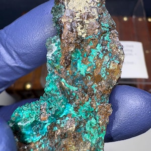 May include: A close-up of a rough, irregular shaped rock with a mix of copper, green, and gold colors. The rock has a rough, textured surface.