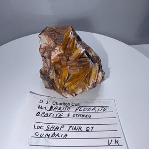 May include: A close-up of a rock sample with orange and brown crystals. The label reads "D.J. Charlton Coll", "Min: Barite Fluorite Apatite + Others", "Loc: Shap Pink QY Cumbria UK".