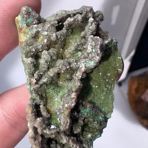 Könnte beinhalten: Nahaufnahme einer Mineralprobe mit rauer, strukturierter Oberfläche. Das Mineral zeigt eine Mischung aus grünen, grauen und orangen Farbtönen. Die grünen Bereiche scheinen die dominierende Farbe zu sein, wobei Grau und Orange Kontraste hinzufügen. Die Probe wird von einer Person gehalten.