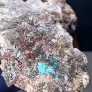 May include: A close-up of a rough rock formation with a mix of colors. The rock has a gray base with patches of reddish-brown and vibrant turquoise. Small flecks of yellow are also visible.