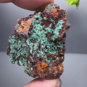 Könnte beinhalten: Nahaufnahme einer Mineralprobe mit einer rauen, dunkelbraunen Matrix, die orangefarbene Bereiche und Ansammlungen kleiner, runder, hellgrüner Formationen aufweist. Die Probe wird zwischen Fingern gehalten.