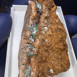 Könnte beinhalten: Ein rauer, brauner Stein mit grünen und weißen Mineralablagerungen. Der Stein hat eine unregelmäßige, zerklüftete Form.