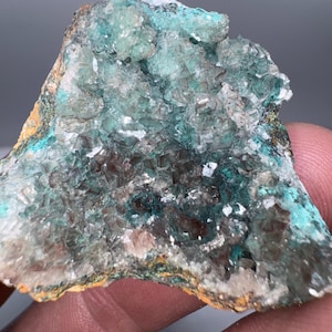 Könnte beinhalten: Nahaufnahme einer Mineralprobe mit einem Cluster aus teal-farbenen Kristallen. Die Kristalle haben ein strukturiertes, kristallines Aussehen mit Andeutungen von Braun und Weiß. Die Ränder der Probe weisen eine Mischung aus orangefarbenen und weißen Farbtönen auf.