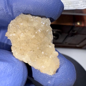 Op de afbeelding: Een close-up van een cluster van kleine, witte, kristallijne formaties. De kristallen zijn geclusterd in een ruwe, onregelmatige vorm en hebben een licht doorschijnende uitstraling.