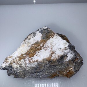 May include: A rough, gray, white, and brown rock with a layered texture. The rock has a white, powdery coating on the surface.