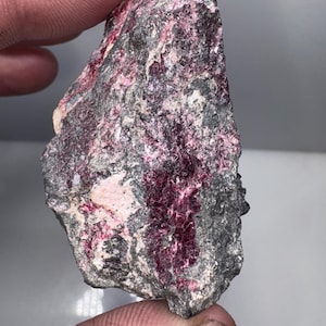 May include: A rough-hewn mineral specimen with a mix of gray and pinkish-red hues. The rock's surface has a textured appearance, with visible crystalline structures and variations in color. The specimen is held between two fingers.