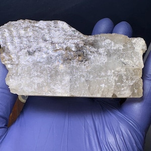 May include: A large, clear, rectangular piece of selenite crystal with a rough, natural texture. The crystal has a slight gray tint and is translucent.