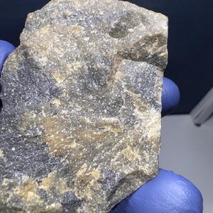 May include: A close-up of a rough, gray and tan rock with a mottled surface. The rock has a layered appearance with darker gray streaks and lighter tan patches.