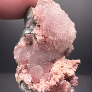 以下が含まれることがあります： ピンク色の鉱物標本のクローズアップ。結晶構造は、ざらざらした質感の表面を持つ、明るいピンク色をしています。鉱物の基部は、濃い緑色と灰色です。