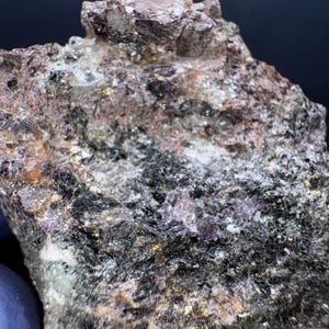 May include: A close-up of a rough, dark grey rock with small, shiny, silver-coloured crystals embedded in it. The rock has a slightly reddish hue in some areas.