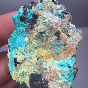 May include: A close-up of a mineral specimen with a rough, textured surface. The stone features a vibrant mix of colors, including turquoise, black, yellow, and brown. The mineral appears to be a natural formation, showcasing a variety of textures and hues.