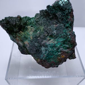 May include: A rough-hewn mineral specimen with a dark green and brown color palette. The surface has a textured, uneven appearance, with areas of deep green and hints of metallic sheen. The mineral sits on a clear acrylic display stand.