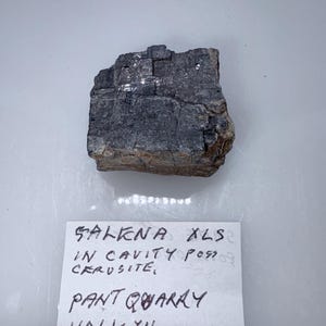 May include: A dark gray, rectangular rock with a white label that reads "GALENA XLS IN CAVITY POSS CERUSSITE, PANT QUARRY HALCYN N. WALES".