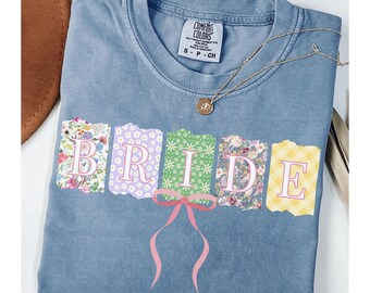 May include: A light blue t-shirt with a "BRIDE" graphic made of colourful patterned fabric squares. The letters are outlined in pink and a pink bow is between the "I" and "D".