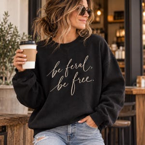 May include: A black long-sleeve crewneck sweatshirt featuring the white cursive text "be feral, be free." across the chest. The garment has a relaxed fit with ribbed cuffs and a ribbed neckline, constructed from a solid black fabric.