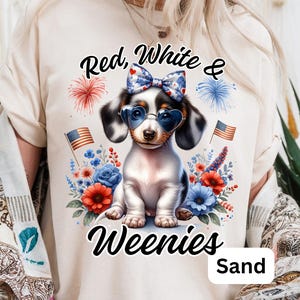 A woman wears a sand-colored t-shirt with a patriotic design featuring a piebald dachshund in heart sunglasses and a bow, surrounded by red, white, and blue flowers, American flags, and fireworks. The text reads “Red, White & Weenies.”