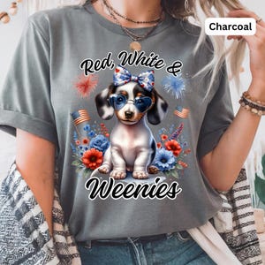 May include: Charcoal-colored t-shirt featuring a graphic of a dachshund wearing heart-shaped sunglasses and a bow. The design includes the text "Red, White & Weenies" with floral and firework accents, and American flags.