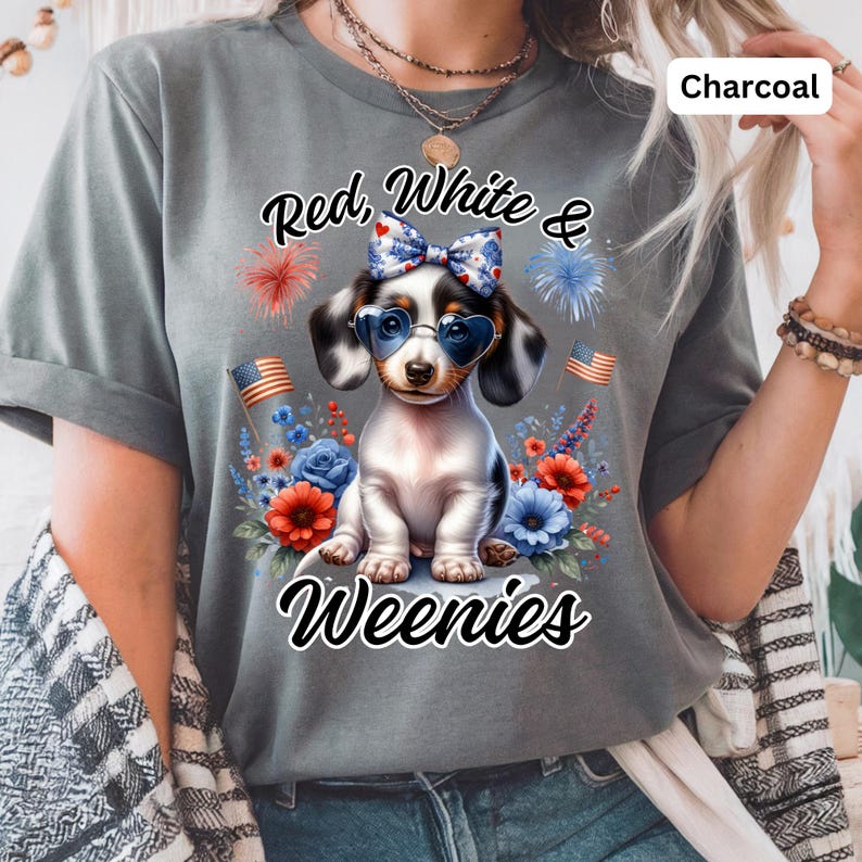 May include: Charcoal-colored t-shirt featuring a graphic of a dachshund wearing heart-shaped sunglasses and a bow. The design includes the text "Red, White & Weenies" with floral and firework accents, and American flags.