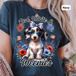 May include: Navy blue t-shirt featuring a dachshund wearing heart-shaped sunglasses and a bow. The shirt has the text "Red, White & Weenies" with fireworks and floral accents. The shirt also has the word "Navy" in the upper right corner.