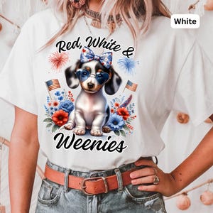 May include: White t-shirt featuring a patriotic dachshund wearing heart-shaped sunglasses and a bow. The shirt has the text "Red, White & Weenies" with floral and firework graphics. The design incorporates red, white, and blue colors.