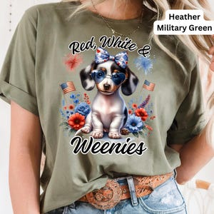 May include: Heather military green t-shirt featuring a patriotic dachshund wearing heart-shaped sunglasses and a bow. The shirt has the text "Red, White & Weenies" with floral and firework accents. Two American flags are also visible.