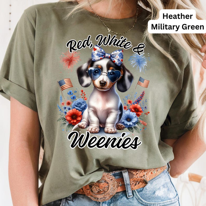 May include: Heather military green t-shirt featuring a patriotic dachshund wearing heart-shaped sunglasses and a bow. The shirt has the text "Red, White & Weenies" with floral and firework accents. Two American flags are also visible.