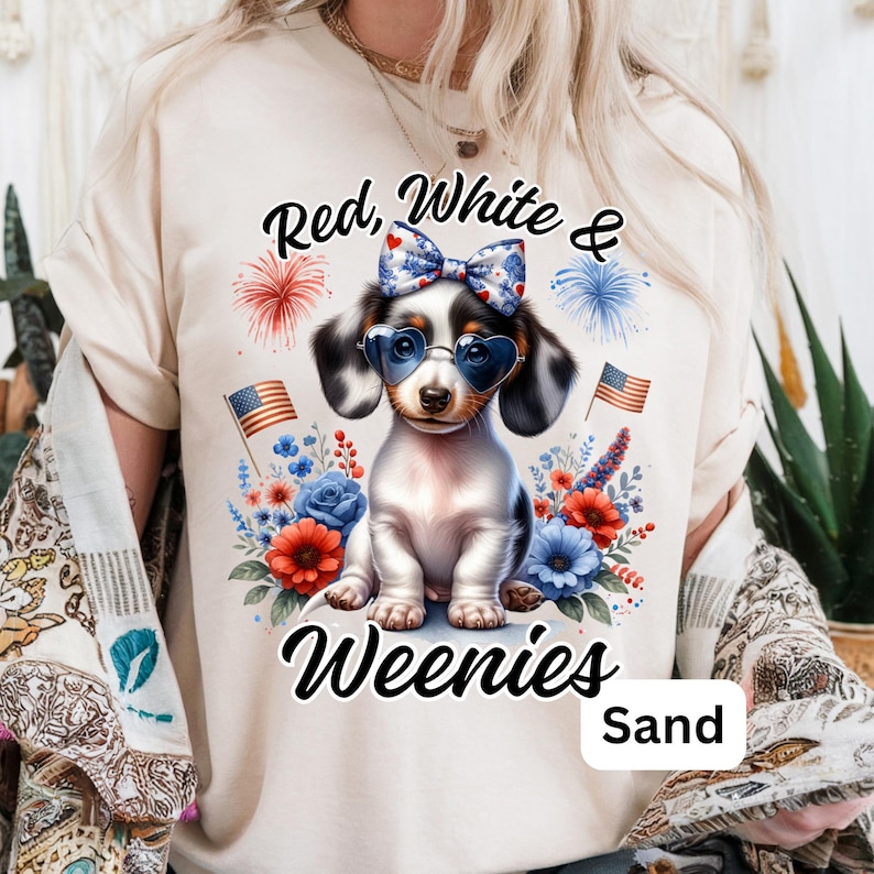 A woman wears a sand-colored t-shirt with a patriotic design featuring a piebald dachshund in heart sunglasses and a bow, surrounded by red, white, and blue flowers, American flags, and fireworks. The text reads “Red, White & Weenies.”
