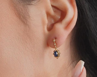 Könnte beinhalten: Nahaufnahme einer Person, die einen goldenen Ohrring mit einem dunkelblauen Edelstein und kleinen weißen Akzenten trägt. Der Ohrring baumelt am Ohr. Die Person hat dunkles Haar und trägt ein schwarzes Oberteil.