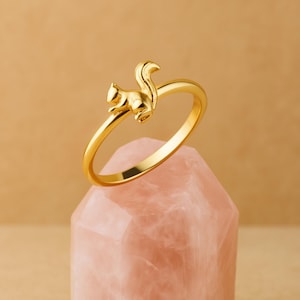 以下が含まれることがあります： 金色のリングで、リスのデザインが施されています。リスはリングのバンドに座っており、ふさふさの尻尾がアーチ状になっています。リングは、暖かくニュートラルな背景に対して、ピンク色のクリスタルに展示されています。