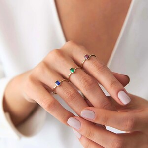 May include: Three gold rings, each with a rectangular gemstone in blue, green, and red. The rings are shown on a hand with manicured nails, against a soft, neutral backdrop. The gemstones are approximately 0.6 cm in length.