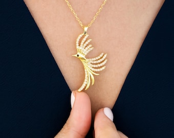 Pode incluir: Um colar com pingente de pássaro dourado com uma corrente delicada. O pingente de pássaro apresenta um design detalhado com pedras claras brilhantes e um pequeno detalhe preto. O colar é exibido em um fundo azul escuro.