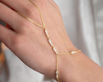 Op de afbeelding: Een delicate gouden handkettingarmband. De armband heeft een fijne ketting met baguette-geslepen heldere stenen en verbindt met een ring. De armband wordt op een hand gedragen.