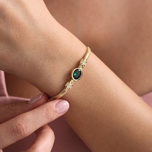 Op de afbeelding: Een gouden armband met een groene ovale edelsteen en kleine heldere stenen.