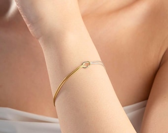 Puede incluir: Un brazalete de oro y plata con un cierre simple de forma ovalada. El brazalete es fino y delicado, y se muestra en la muñeca de una persona.