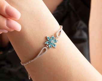 Può includere: Bracciale in argento con un design a fiocco di neve, con pietre preziose blu chiaro e una pietra centrale trasparente. Il bracciale è indossato su un polso, mettendo in risalto la sua delicata catena e gli elementi decorativi.