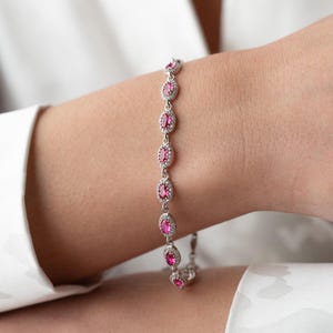 May include: A silver bracelet featuring oval-shaped, bright pink gemstones surrounded by small, clear stones. The bracelet is displayed on a wrist, showcasing its delicate design and sparkling details. A classic piece of jewelry.