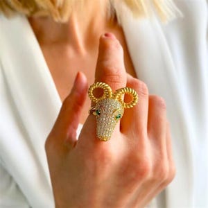 May include: A gold ring featuring a detailed ram's head design. The ram's head is adorned with clear rhinestones and has green gemstone eyes.