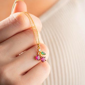 May include: A gold-colored necklace featuring a cherry pendant. The pendant has two red gemstone cherries, a green gemstone leaf, and a small white gemstone. The necklace is held in a hand.