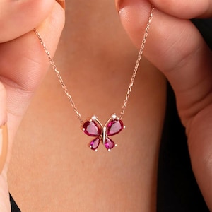 Peut inclure: Un collier pendentif papillon en or rose. Le motif papillon présente des pierres précieuses roses vibrantes et de petits accents blancs. Le collier est tenu par les mains d'une personne, mettant en valeur la chaîne délicate et les détails du pendentif.