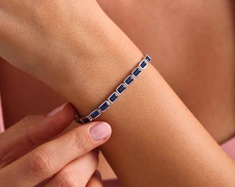 May include: A silver bracelet with rectangular, dark blue gemstones. The bracelet is worn on a wrist, showcasing the design and color contrast. The gemstones are evenly spaced, creating a sleek and elegant look. The bracelet is a piece of jewelry.