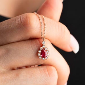 Könnte beinhalten: Eine zarte goldene Halskette mit einem tropfenförmigen Anhänger, der einen großen roten Rubin umrahmt, der von kleineren Diamanten umgeben ist.