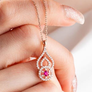 Puede incluir: Un collar con colgante de oro rosa con un diseño de lágrima. El colgante está adornado con pequeñas piedras preciosas transparentes y una piedra preciosa roja redonda en el centro. El collar lo sostiene una persona.