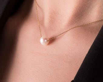 May include: A delicate gold chain necklace featuring a single, lustrous white pearl pendant. The pearl is round and centrally positioned, suspended from a fine gold chain. The necklace is shown against a dark background.
