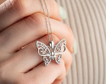 May include: A silver butterfly pendant necklace. The butterfly pendant is encrusted with small, sparkling stones and features an openwork design. The necklace is held by a person.