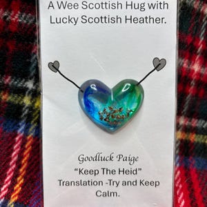 May include: A small heart-shaped glass ornament with blue and green swirls and a small amount of dried heather inside. The ornament is attached to a card that reads "A Wee Scottish Hug with Lucky Scottish Heather. Goodluck Paige "Keep The Heid" Translation -Try and Keep Calm."