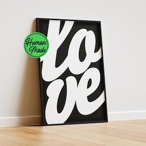 Op de afbeelding: Een rechthoekige zwarte lijst met een witte poster waarop het woord "love" in grote, dikke cursieve letters staat. Een ronde groene sticker met de tekst "Human Made" is geplaatst op de linkerbovenhoek van de lijst.