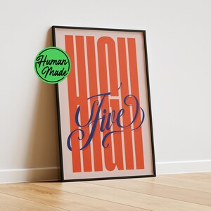 Könnte beinhalten: Ein gerahmter vertikaler Wandkunstdruck mit hellrosa Hintergrund und kräftigen, hohen, orangefarbenen vertikalen Streifen. In der Mitte steht der Text "High Five" in einer dunkelblauen, fließenden Schreibschrift. Ein grünes kreisförmiges Logo zeigt "Human Made".