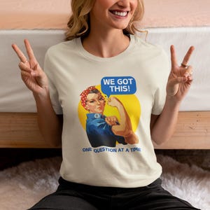 May include: A white t-shirt with a vintage-style illustration of a woman with her fist raised. The text "We Got This!" is in a blue speech bubble above her head. The text "One Question At A Time" is below the illustration.