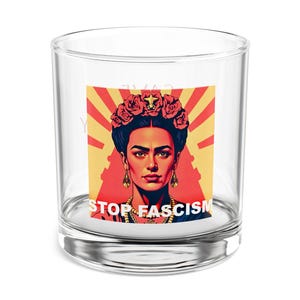 May include: A clear glass votive candle holder with a Frida Kahlo graphic, floral crown, and the text "STOP FASCISM". The background is yellow with red rays. A decorative item.