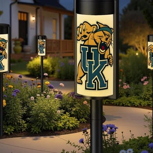 May include: Black outdoor pathway lights with a University of Kentucky Wildcats logo. The lights have a cylindrical design with a black top and base, and a glowing panel displaying the team logo. The lights are placed along a garden path.